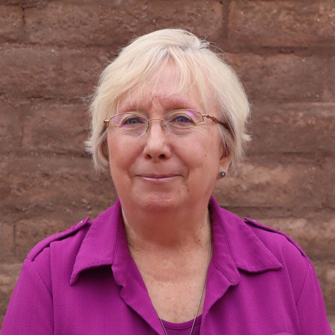 A woman in purple shirt and glasses standing next to brick wall.